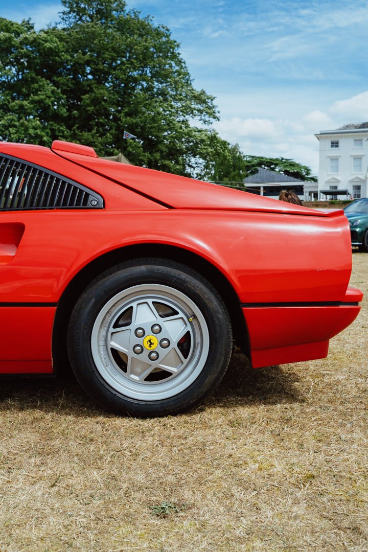 Close-up of the rear side of a red Ferrari sports car parked on grass, with a white house and trees in the background.