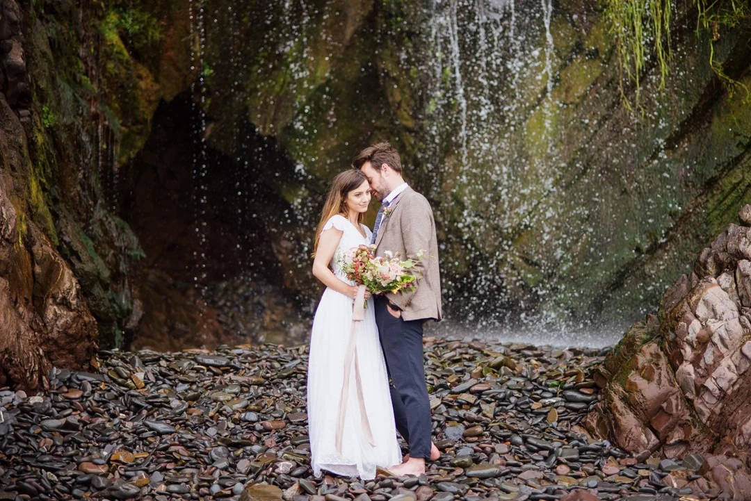 bride and groom stood near large waterfall on stoney beach, the bride is holding red and green flowers