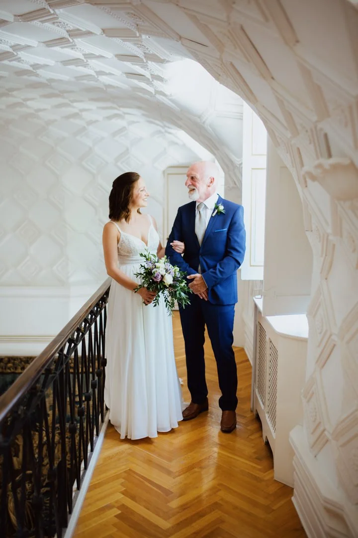 A bride and an older man, possibly her father, standing together indoors, smiling and looking at each other. The bride is holding a bouquet of flowers, wearing a white wedding dress, and the man is in a blue suit with a tie. The setting has ornate wh