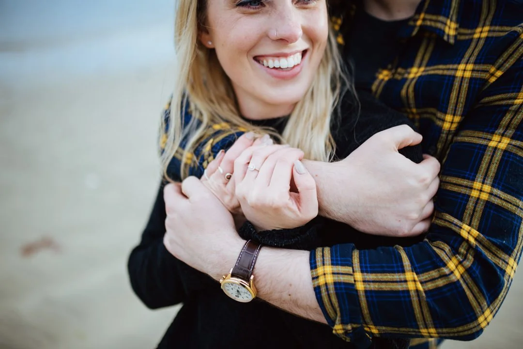A woman smiling as she embraces someone, with her arms crossed around them. She has blonde hair, wears a black top, and a watch on her wrist. The person she is hugging is wearing a plaid shirt.