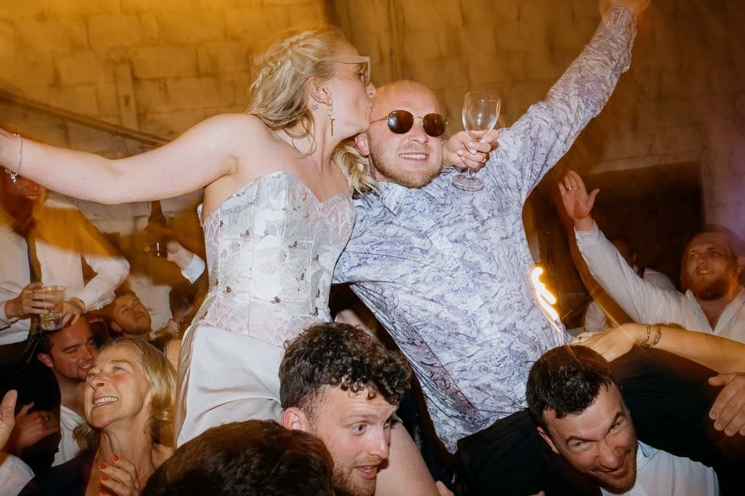 People celebrating at a party with a woman standing on a man's shoulders, kissing him on the cheek while he holds a glass of wine, surrounded by smiling guests.