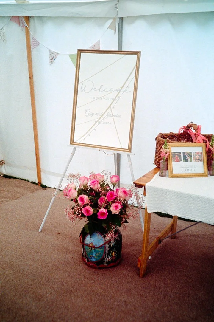 Wedding welcome sign with pink roses in a vase at a reception table with cards and decorations.