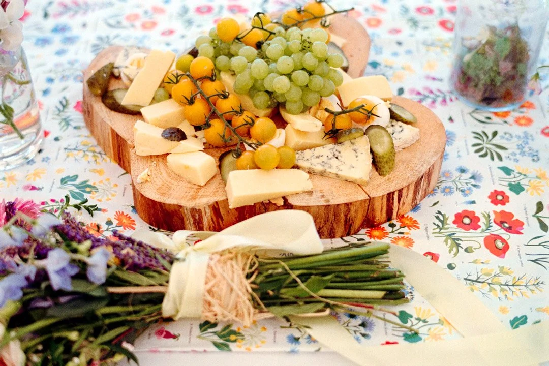 A wooden cheese board with various cheeses, grapes, cherry tomatoes, and pickles on a floral tablecloth, with a bouquet of flowers in the foreground.