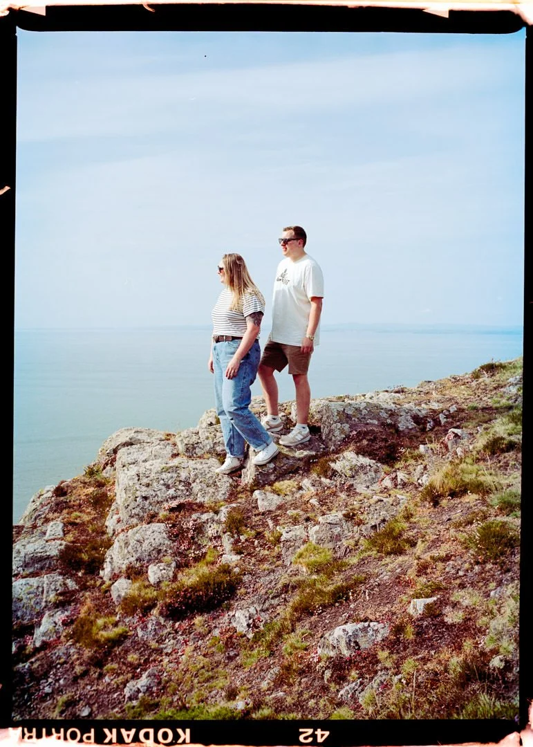 A man and woman standing on rocky terrain near the water's edge, overlooking a body of water under a partly cloudy sky.