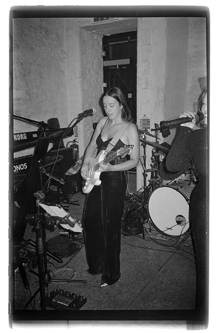 A woman playing electric guitar and singing into a microphone during a live music performance in a room with stone walls.