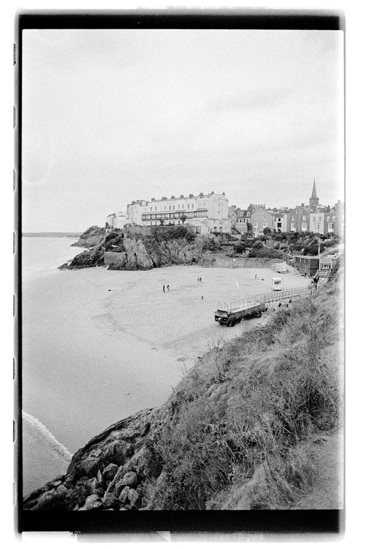 A black and white photo of a coastal scene with a sandy beach, rocky cliffs, and buildings in the background, including a tall church steeple.