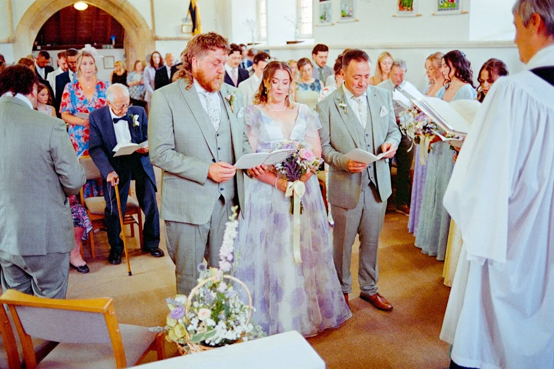 A wedding ceremony inside a church with the bride and groom standing at the altar, holding papers, surrounded by guests, and officiated by a priest.