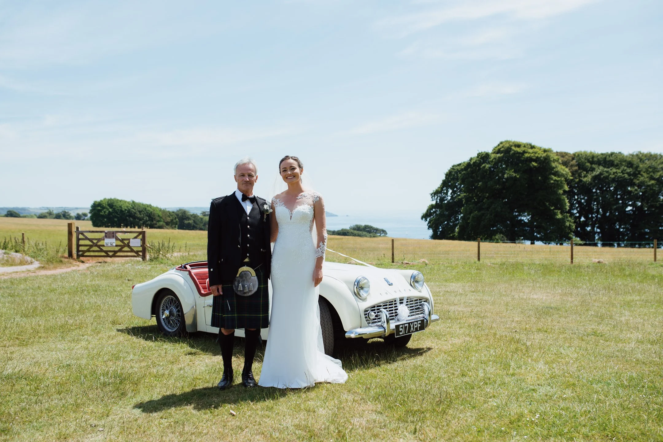 A bride in a white wedding dress and an older man in a kilt and black tuxedo standing next to a vintage white convertible car on a grassy field with trees and a blue sky in the background.