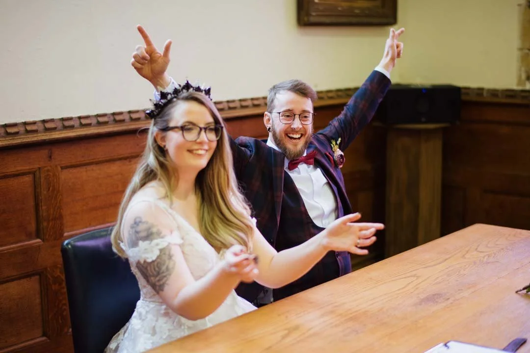 bride and groom sat at large wooden table smiling and waving hands in the air