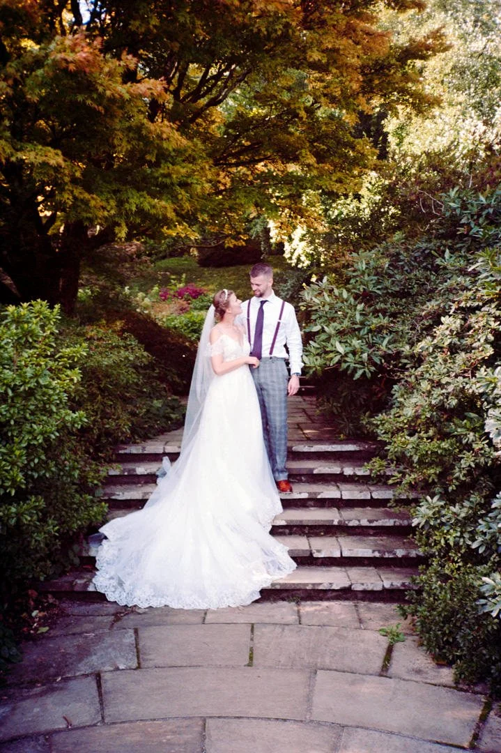 A bride and groom standing on stone steps outdoors, dressed in wedding attire, sharing a tender moment among lush greenery and trees.