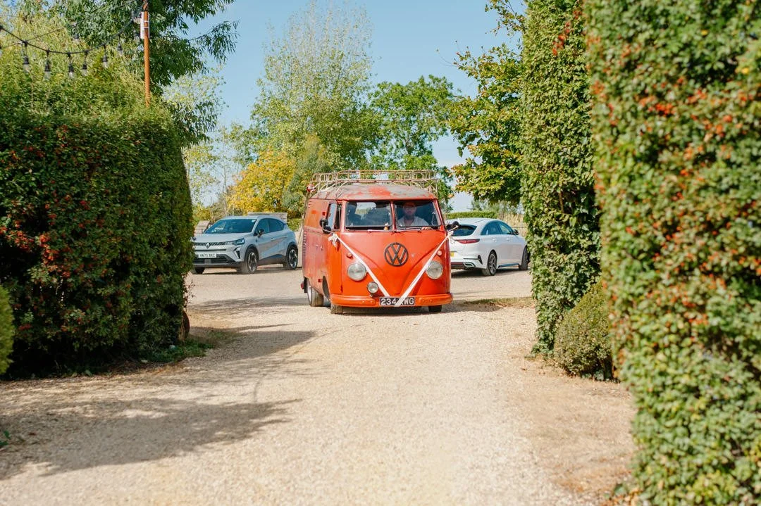 A red vintage Volkswagen van with a large VW logo on the front, driving down a gravel path surrounded by green hedges and trees, with two modern cars behind it.