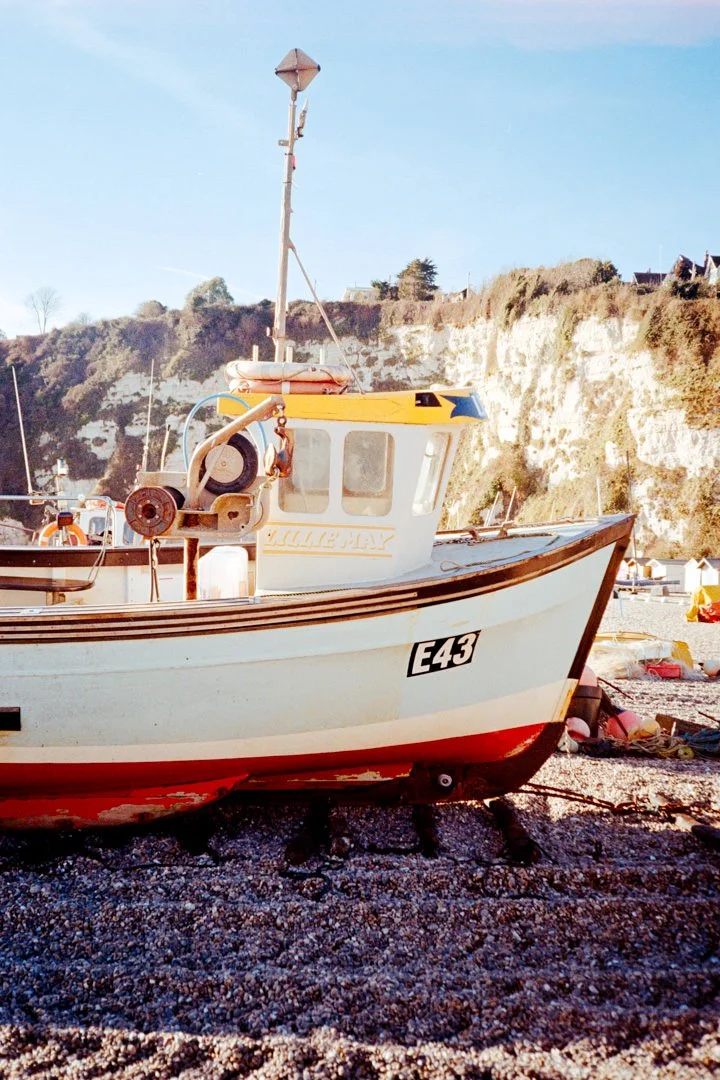 A small fishing boat resting on a beach with rocky terrain, hillside with houses in the background, and a clear blue sky.