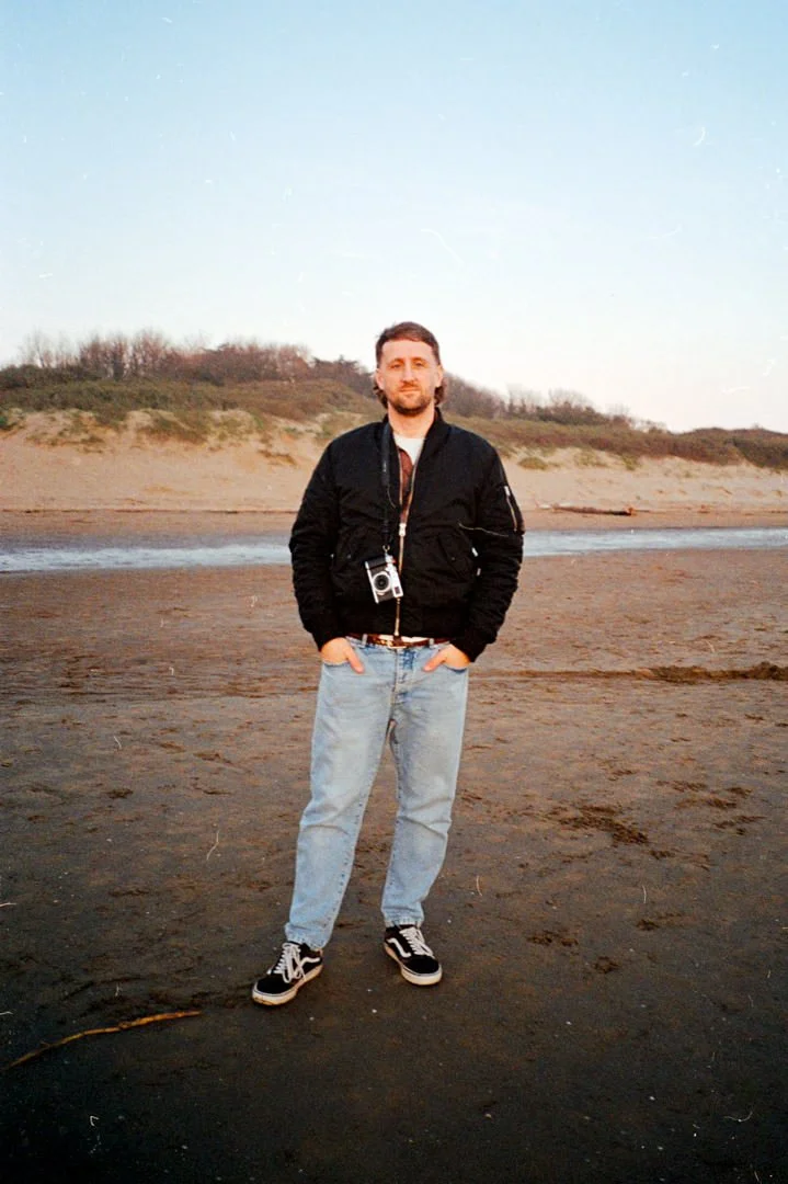 man waring black bomber jacket holding film camera on beach