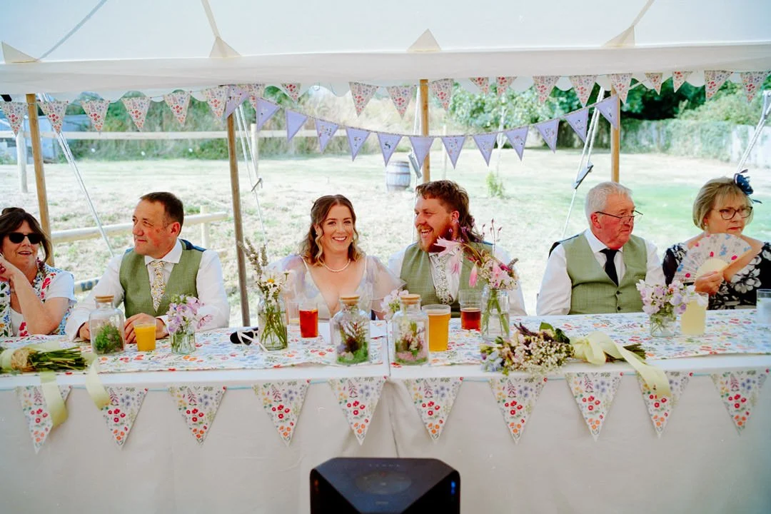 Wedding reception table decorated with floral arrangements and colorful drinks, under a tent with bunting decorations, with happy guests including a bride and groom smiling at each other.
