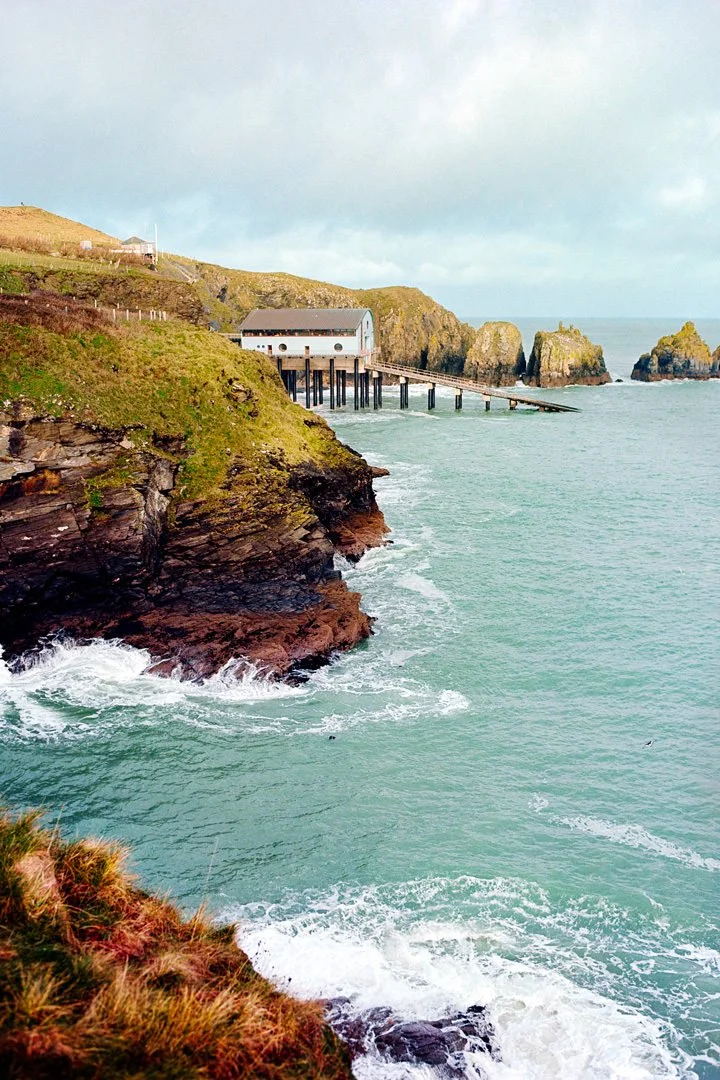 Cliffside view of a coastal pier leading to a white building over the ocean, with rocky formations in the background and green grassy land in the foreground.