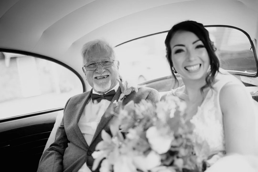 black and white photo of a wedding bride sat next to her father. they are sat on the back seat of an old car