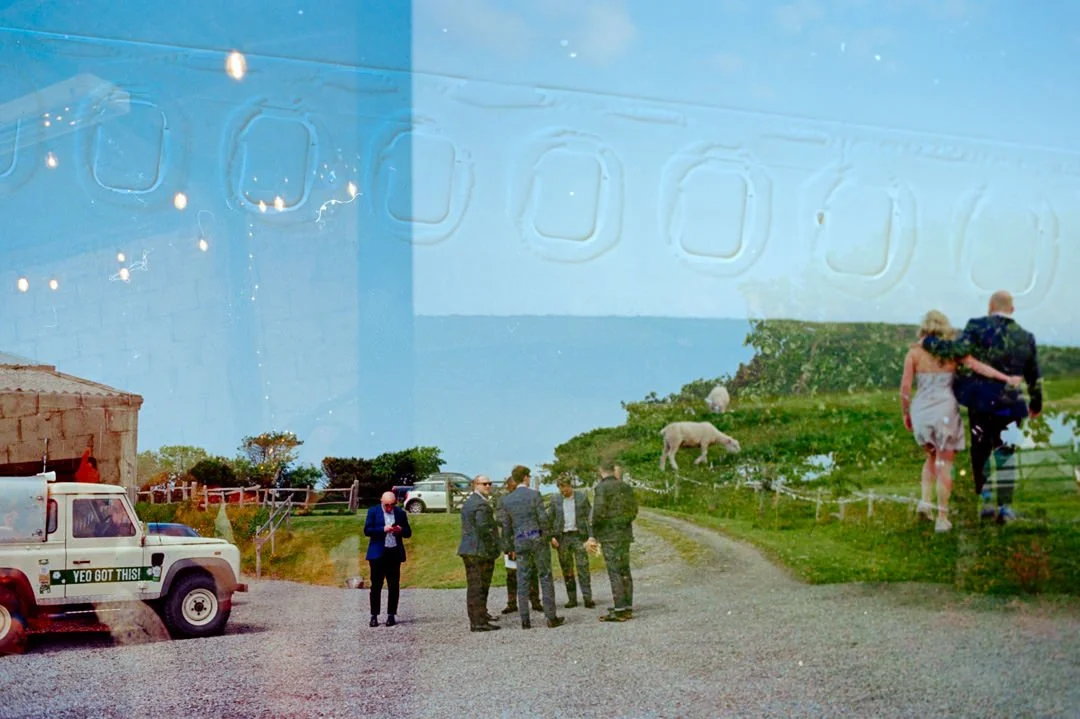 A reflection on a window showing a rural scene with a group of five men in suits talking, a woman and man walking together, a cow grazing, a white truck with a sticker that says 'YEO GOT THIS!', and green fields under a blue sky.