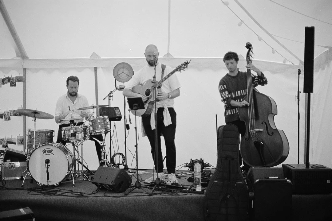 Three musicians performing on a stage inside a tent: a drummer on the left, a guitarist in the center, and a double bassist on the right.