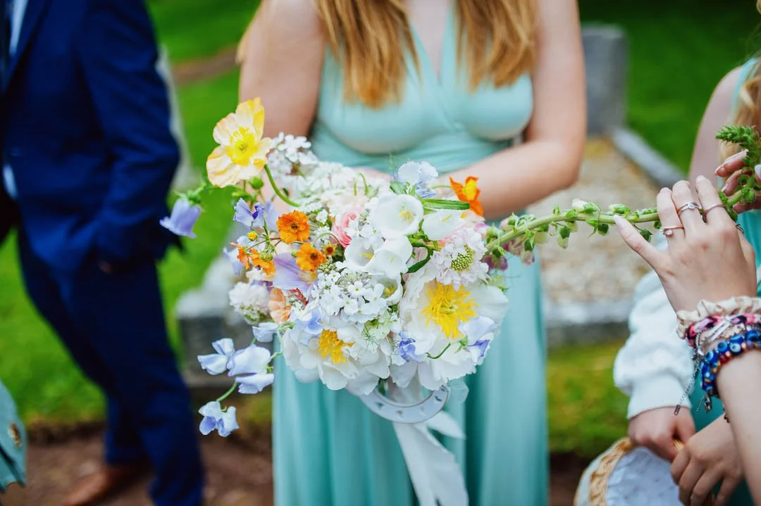 A woman in a mint green dress holding a bouquet of colorful flowers, including white, yellow, orange, and purple blossoms, at an outdoor event.