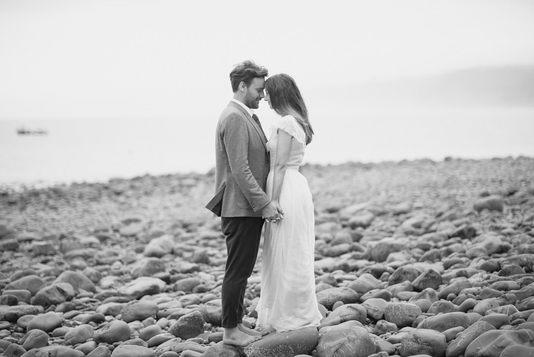 bride and groom stood on beach holding hands with boat in the background
