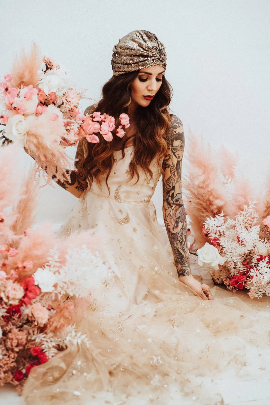 A woman with long wavy brown hair and tattoos on her arms wearing a vintage-style cream dress with moon and star embroidery, sitting among pink and white flowers, holding a bouquet of pink flowers, and wearing a sparkly gold headscarf.