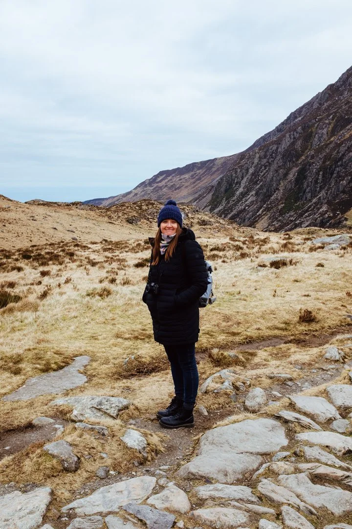 A woman in a black jacket, blue beanie, and hiking boots stands on a rocky trail in a mountainous landscape with dry grass and steep hills in the background.