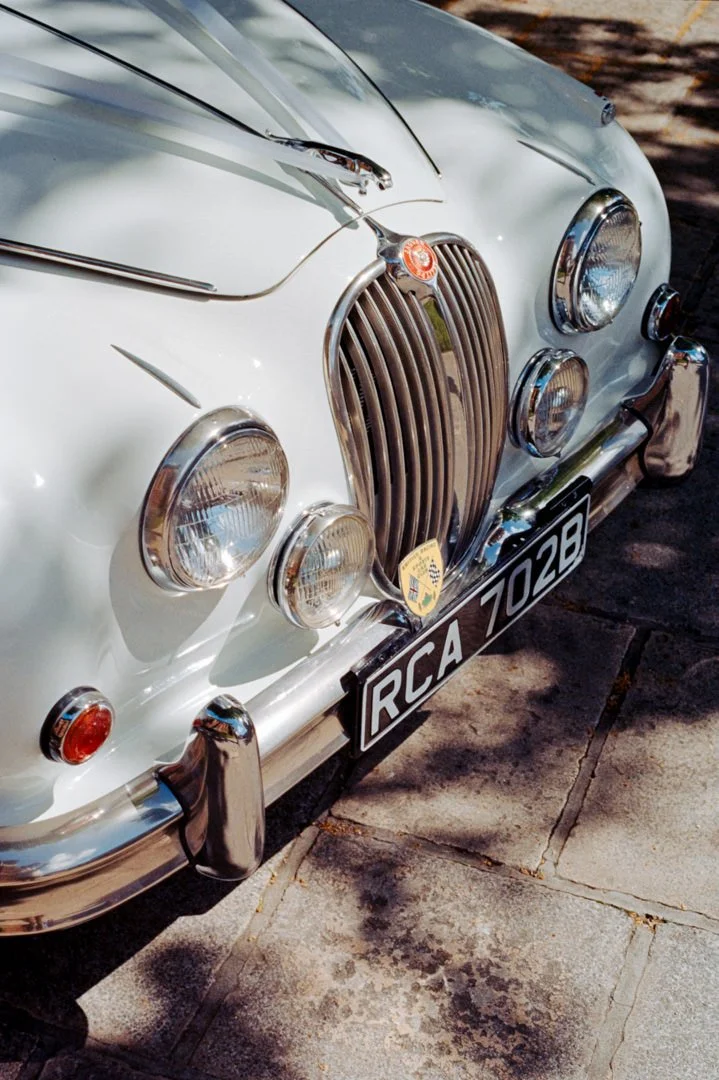 Close-up of the front of a white vintage Jaguar car, showing the grille, four headlights, chrome bumper, and a black license plate with the text "RCA 7028."