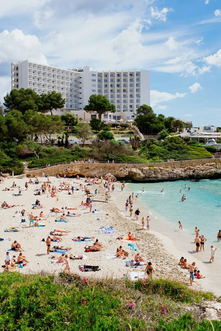 People sunbathing and swimming at a crowded beach, with a large white hotel and green trees in the background under a partly cloudy sky.