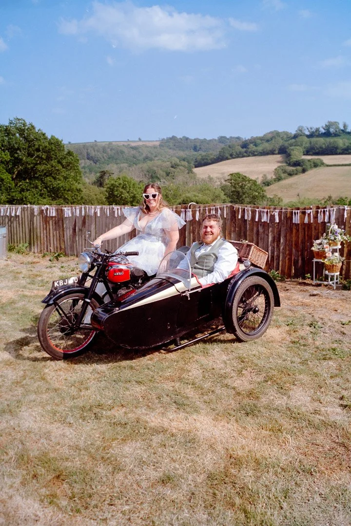 Woman in a wedding dress and sunglasses sitting on a vintage motorcycle with a sidecar, with a man in a vest and dress shirt sitting in the sidecar, outdoors in a grassy area with a wooden fence and rolling hills in the background.