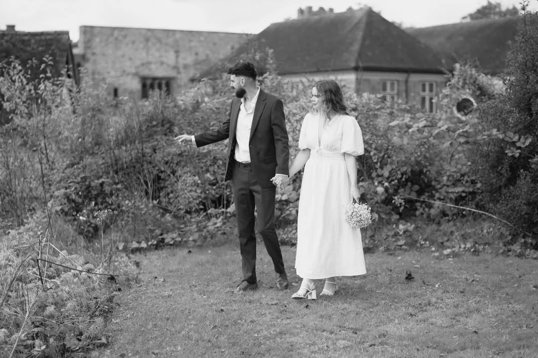 bride and groom walking in garden holding hands, the bride is holding flowers and looking up at the sky