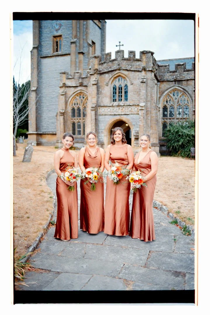 Four women in matching bronze dresses holding bouquets, standing in front of a historic stone church with stained glass windows.