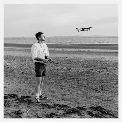 man in white t shirt and black shorts on beach flying a drone, he has a controller in his hand as is looking up
