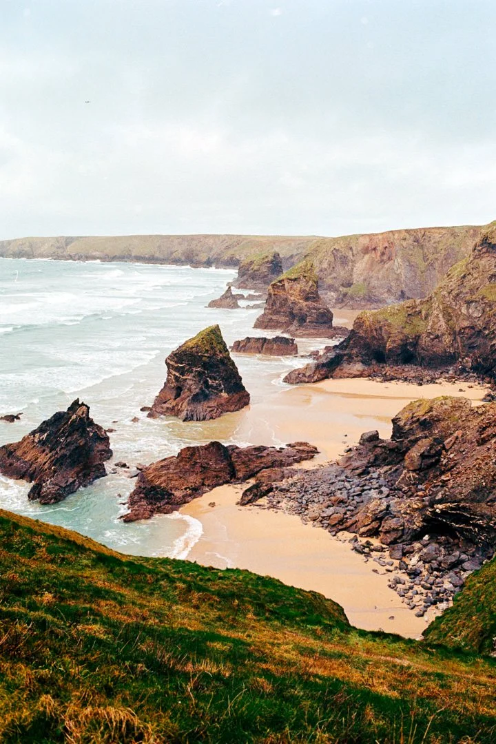 Coastal cliffs with rocky formations and sandy beach under cloudy sky, viewed from an elevated grassy area.