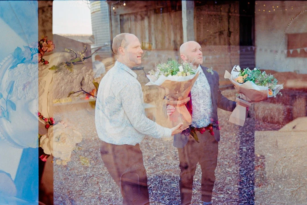 Two men holding bouquets of flowers, seen through a glass window, during a daytime outdoor event.