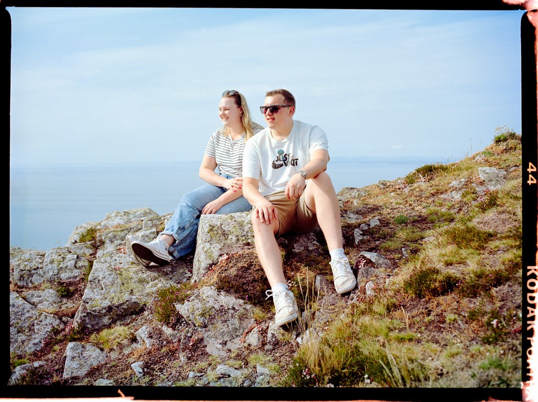 A man and a woman sitting on rocks on a hillside near the coast, smiling and enjoying the sunny day with the ocean in the background.