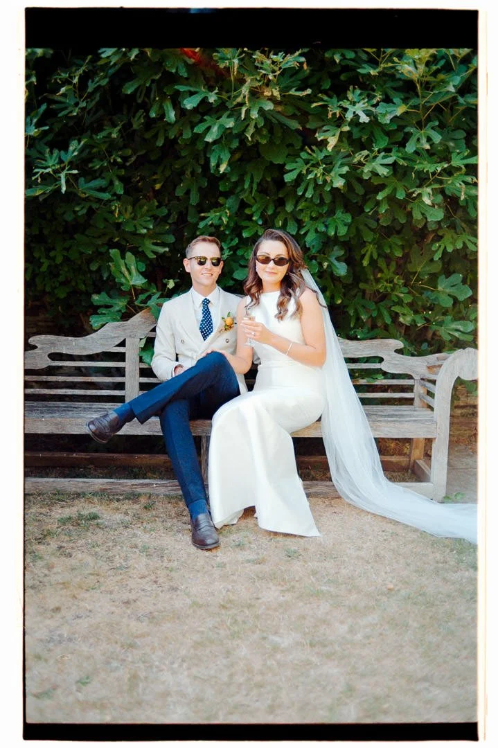 A bride and groom sitting on a wooden bench outdoors, with large green leafy bushes in the background. The bride is wearing a white wedding dress with a veil and holding a glass, while the groom is dressed in a light-colored suit with dark pants, a p