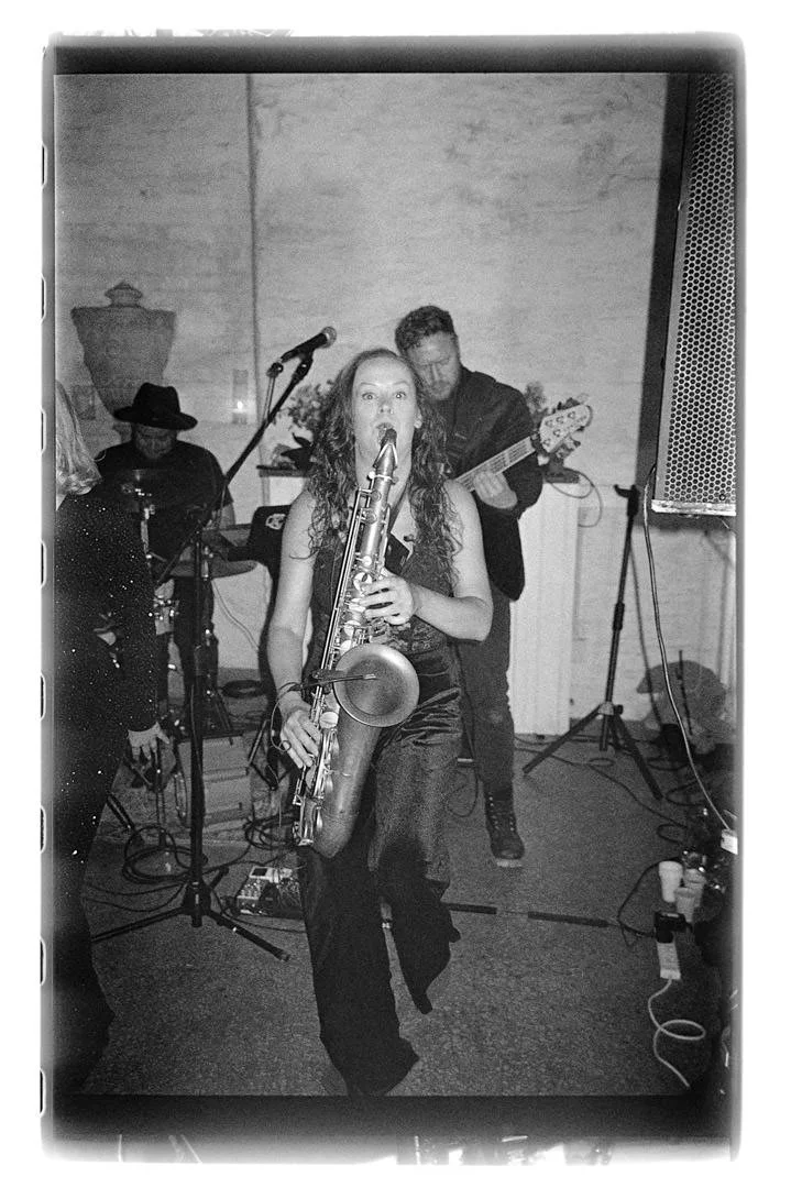 Black and white photo of a woman playing a saxophone and singing into a microphone, with two men playing guitar and drums in the background.