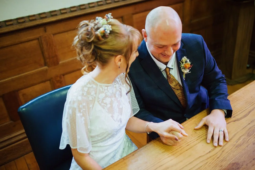 bride and groom sat at table holding hands, they are looking at wedding rings on their hands