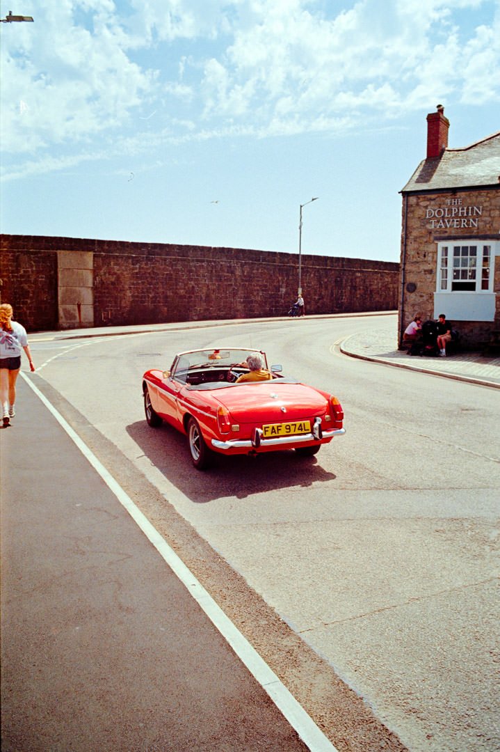 A bright red vintage convertible car with yellow license plate 'FAF 974L' parked on a street near a stone building with a sign that reads 'The Dolphin Tavern.' There are a few people around, one woman walking on the sidewalk, another sitting on a ben