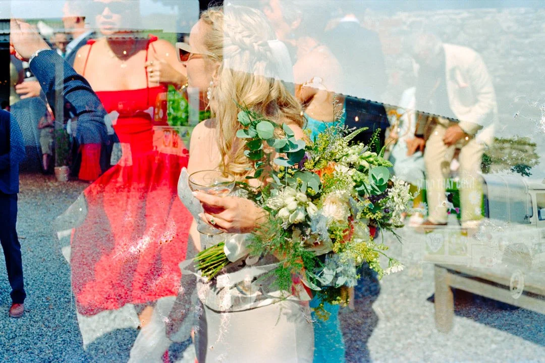 A woman holding a large bouquet of white and green flowers at an outdoor event or wedding, with multiple people and a vehicle visible in the background.