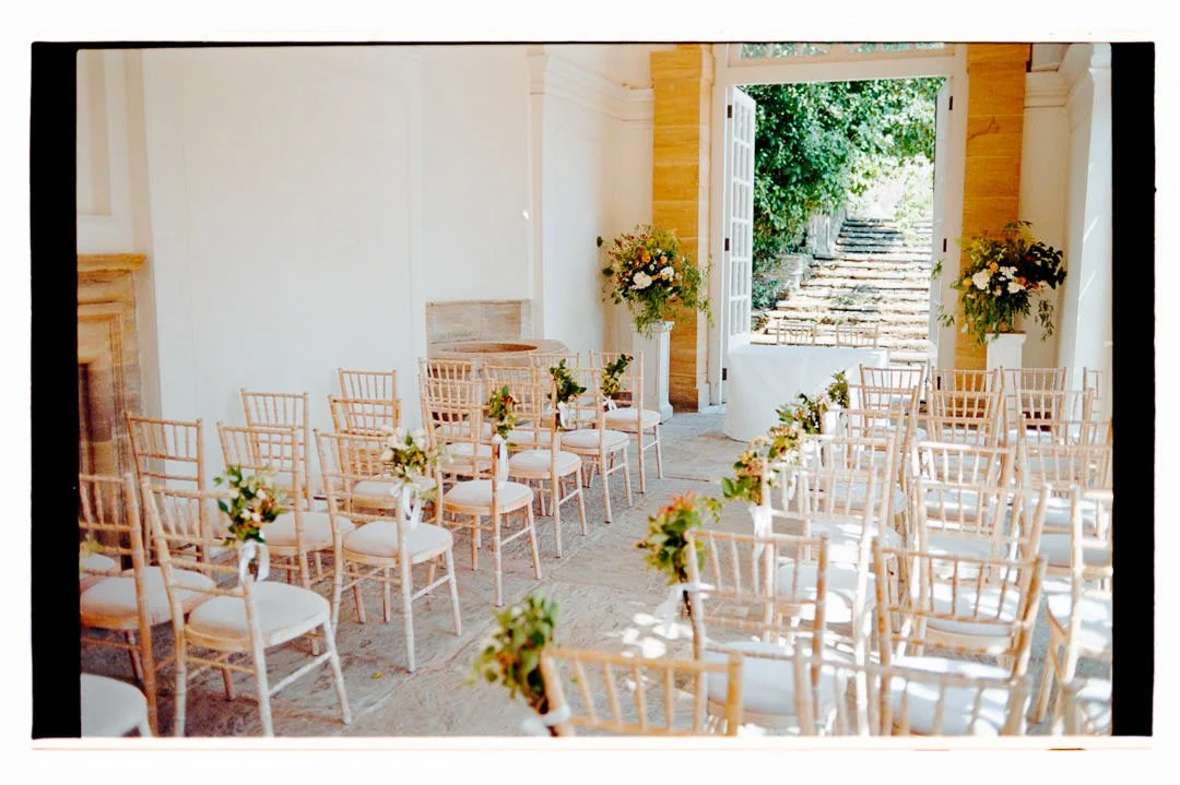 Interior of a wedding ceremony setup with rows of white chairs decorated with floral arrangements, open double doors leading outside to stone steps, and lush green foliage visible
