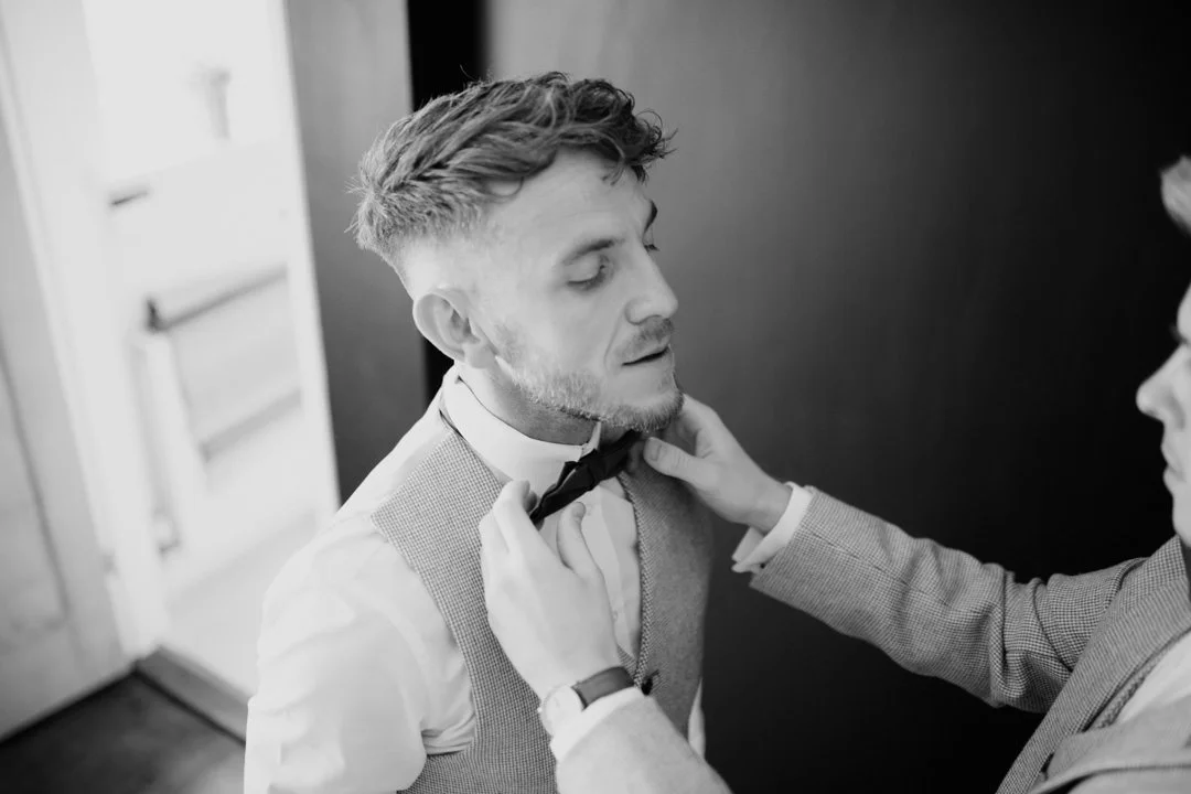 man with short wavy hair putting on dickie bow tie, he is waring a grey waistcoat