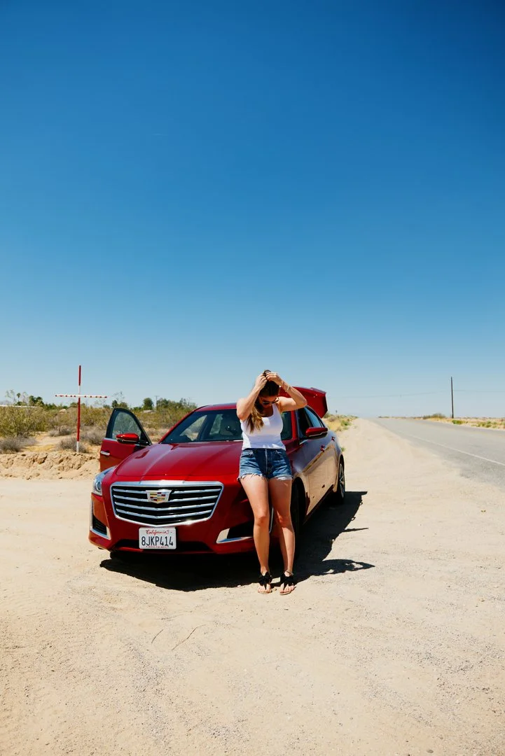 A woman standing in front of a red Cadillac with its door open on a deserted road in a desert landscape, adjusting her hair under a clear blue sky.