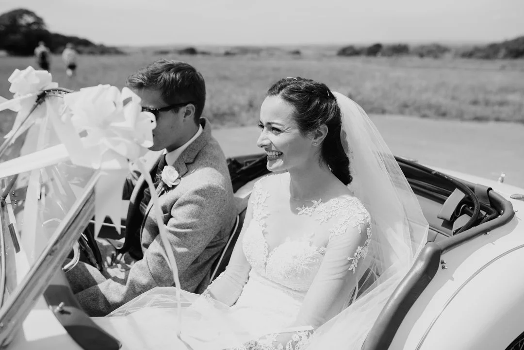 Black and white photo of a smiling bride and a man in a convertible car on a golf course, with open field and some people in the background.
