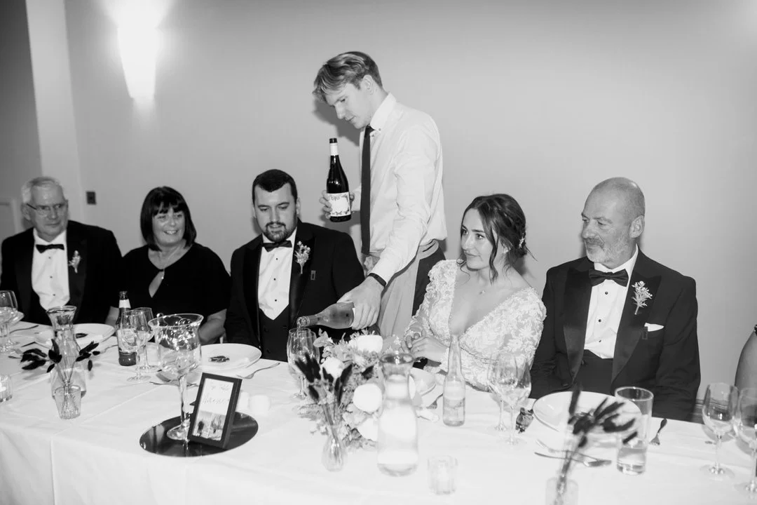 Black and white photo of a wedding reception with six people sitting at a table, and a young man pouring champagne for the bride, who is in a white lace dress, flanked by her groom and family members.