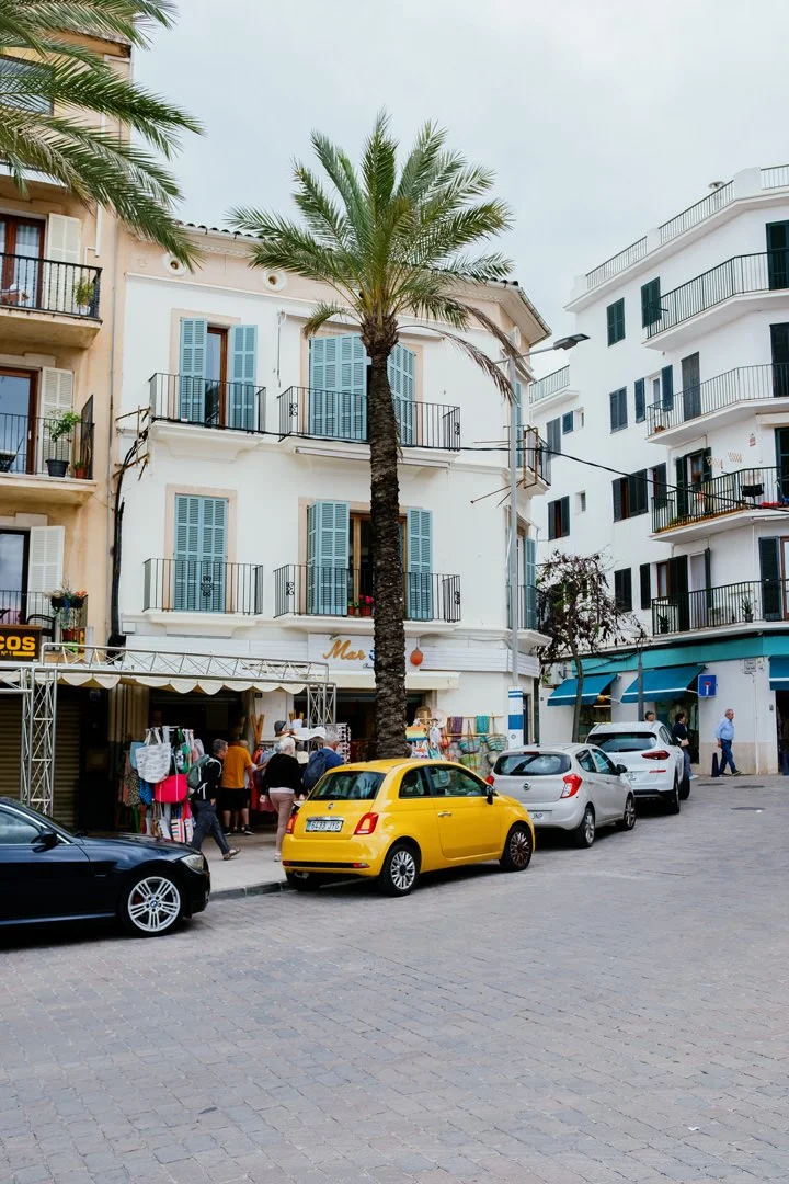A street scene featuring colorful buildings with balconies, a tall palm tree, parked cars including a yellow one, and pedestrians near a small shop.