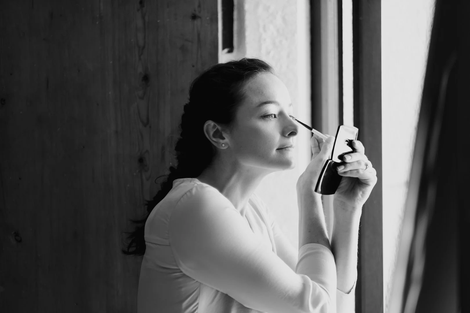 Black and white photo of a woman applying mascara while looking in a mirror near a window.
