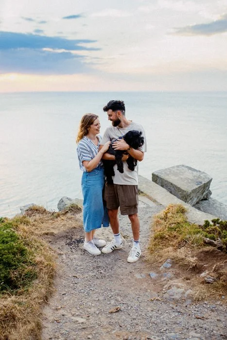 A couple and a dog standing on a rocky path near the ocean at sunset.