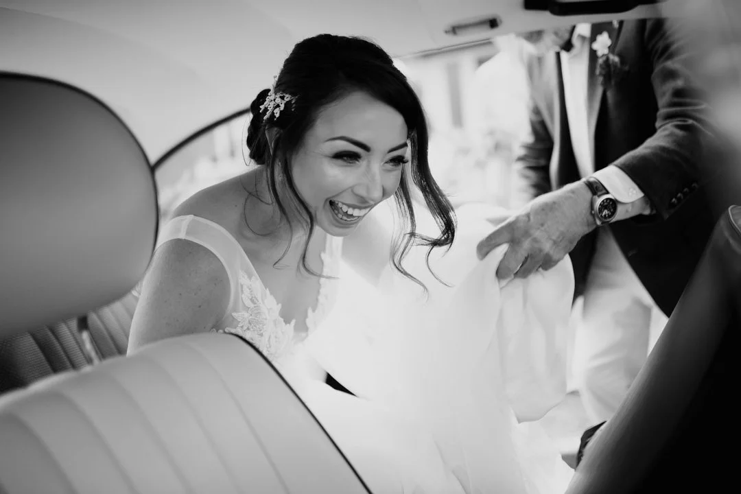 bride waring white dress getting a car. she is smiling looking out of the window