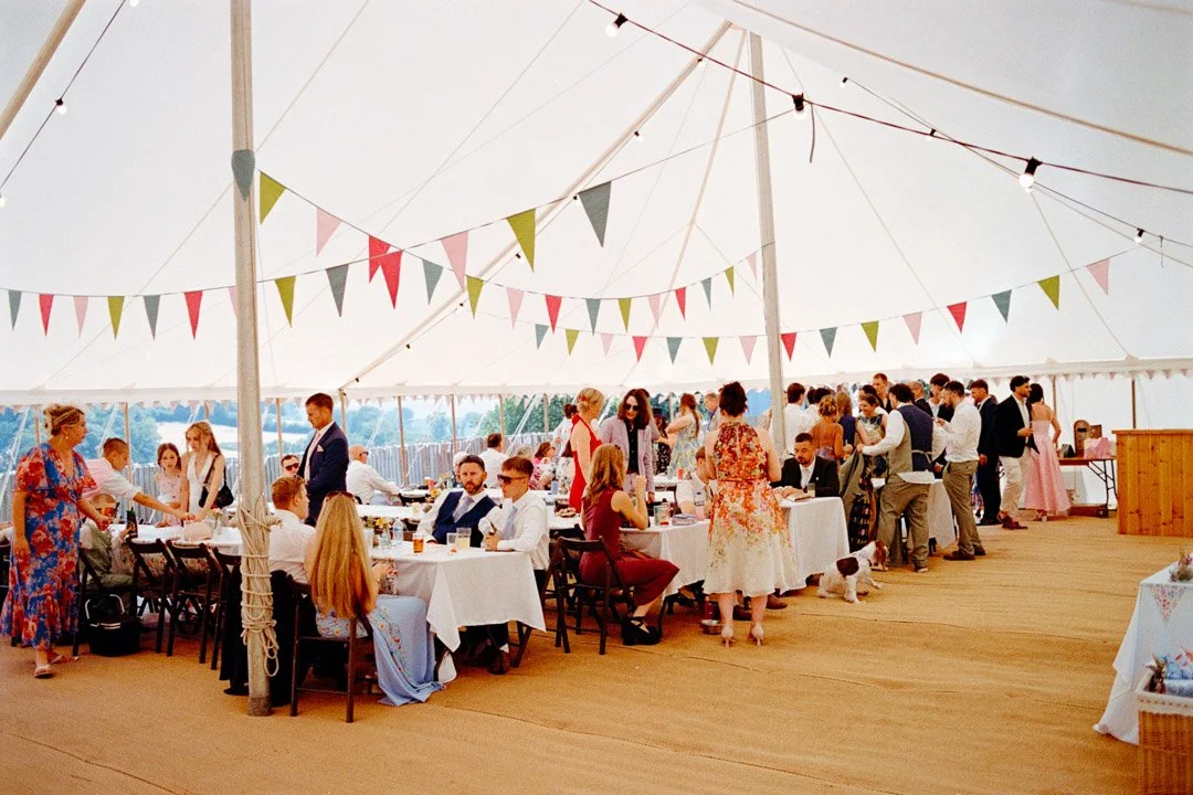 People gathered at a large wedding reception inside a decorated white tent with bunting, tables set for dining, and guests mingling.